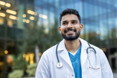 Beyaz önlük ve steteskop giyen kendine güvenen bir tıp uzmanı kameraya bakmadan gülümsüyor ve modern hastane binasının önünde duruyor.