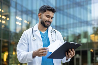 Modern bir hastanenin dışında hasta çizelgelerini inceleyen kendinden emin genç bir erkek doktor, hasta bakımı ve analizi yaparken sağlık uzmanlığı ve profesyonelliği somutlaştırıyor.