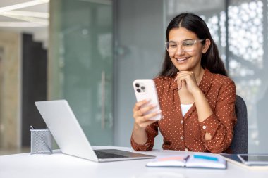 Akıllı telefonu kullanırken gülümseyen mutlu iş kadını ve modern ofisindeki bilgisayarında çalışırken, kariyerindeki teknoloji ve iletişim karışımının keyfini çıkarıyor.