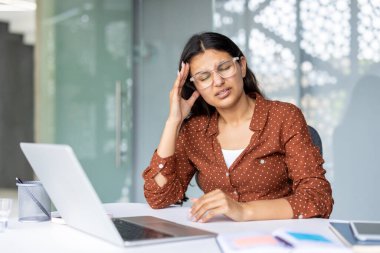 Genç iş kadınının ofiste dizüstü bilgisayarla çalışırken başı ağrıyor. Elleriyle kafasına dokunuyor. Ağrı, migren, stres, aşırı çalışma.