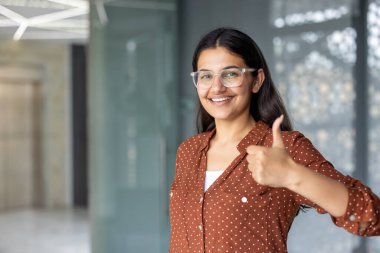 Gözlüklü genç Hintli yönetici gülümsüyor ve başparmak işareti yapıyor. Modern ofis binasında duruyor. Onayını ve memnuniyetini ifade ediyor.