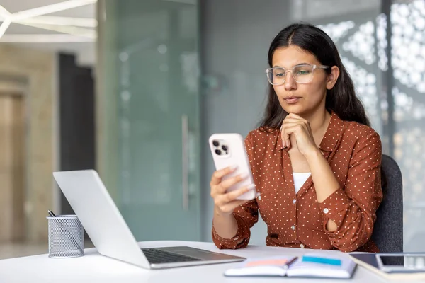 İş yerinde dizüstü bilgisayarla çalışırken cep telefonundan mesaj okuyan genç profesyonel bir kadın, cep telefonunda iş uygulaması kullanan konsantre bir kadın.