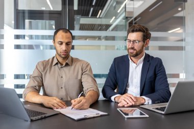 İşadamı dikkatle bir sözleşmeyi inceler ve imzalarken, menajeri de çağdaş ofis ortamında etkin iş akışı için dizüstü bilgisayar ve tabletler kullanır ve rehberlik eder.