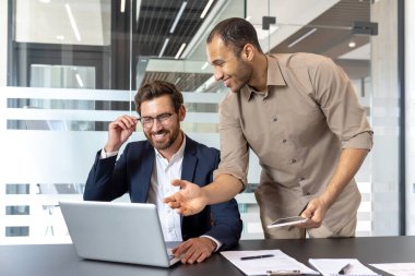 İki neşeli iş adamı, modern ofis ortamında bir dizüstü bilgisayar ve tablet kullanarak takım çalışmalarını ve üretkenliklerini gösteren bir proje üzerinde işbirliği yapıyorlar.