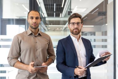 İki işadamı modern bir ofis koridorunda duruyorlar, biri dijital tablet tutuyor, diğeri ise elinde belge ve kalemle birlikte bir pano tutuyor, bu da ortak bir çalışma ortamı olduğunu gösteriyor.