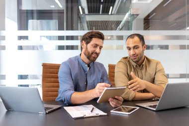 İki gülümseyen erkek yönetici, bir proje üzerinde işbirliği yaparak ve bilgisayarlarla ve tabletlerle çevrili modern ofis ortamında notları tartışarak verimli bir iş toplantısı yapıyorlar.