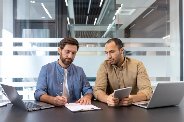 Bir proje üzerinde işbirliği yapan iki işadamı, biri belgede yazarken diğeri ise modern bir ofiste dijital tablet, takım çalışması ve teknolojiyi somutlaştırıyor.