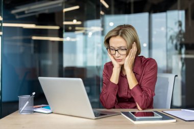 Stresli iş kadını iki eliyle yüzüne dokunuyor, modern bir ofiste masa başında çalışırken bilgisayar ekranına bakıyor, iş baskıları ve teslim tarihleriyle boğuşuyor.