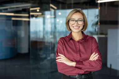 İş kadını kollarını kavuşturarak gülümsüyor, modern ofis ortamında güven ve profesyonellik yayıyor, şirket dünyasında liderlik ve başarı sergiliyor.