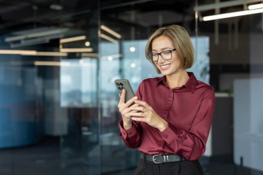 Gözlük takan ve şık kıyafetler giyen mutlu iş kadını modern ofiste akıllı telefon kullanıyor, internet iletişimi ve iş için mobil teknolojinin keyfini çıkarıyor