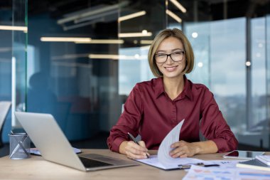 Belgelerle çalışırken gülümseyen ve modern ofiste cam bölmeli dizüstü bilgisayar kullanan mutlu iş kadını verimli iş gününün tadını çıkarıyor.