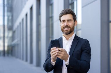 Mutlu işadamı ofis binasının önünde akıllı telefon kullanırken gülümsüyor, internet iletişiminden, başarılı anlaşmadan veya mobil uygulamadan keyif alıyor