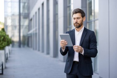 Modern ofis binasının dışında yürürken dijital tablet kullanan kendine güvenen işadamı, şirket ortamında bağlantı, hareketlilik ve verimliliği temsil ediyor.