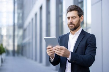 Modern ofis binasının önünde dikilirken dijital tablet kullanan kendine güvenen işadamı, mobil teknolojiyi, iş iletişimini ve modern iş yerini temsil ediyor