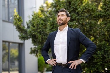 Takım elbiseli bir işadamı, elleri kalçasında dışarıda duruyor, yüzünde kendine güvenen bir ifadeyle, ağaçlarla ve arka planda bir binayla yukarı bakıyor.