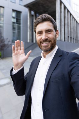 Web kamera görüntüsü, akıllı telefon kamerasına bakıp el sallayan ve gülümseyen bir adam. İş adamı şehirde dolaşıyor ve arkadaşlarıyla internetten konuşuyor..