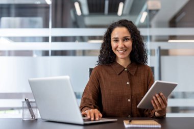 Profesyonel iş kadını kameraya gülümsüyor, aktif olarak dizüstü bilgisayarla çalışıyor ve elinde dijital tablet tutuyor, modern bir ofis ortamında masasında oturuyor.