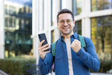 Gözlüklü ve sırt çantalı genç adam dışarıda başarıyı kutluyor, gülümsüyor ve akıllı telefonu modern bir kampüs binasının önünde tutuyor, neşeli ve heyecanlı bir ifade.