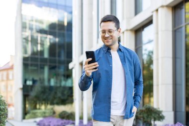 Gözlüklü ve günlük kıyafetlerle gezen genç adam, şehir merkezinde yürürken gülümsüyor, elinde akıllı telefon, mesajlar ve uygulamalar tutuyor. Gündüz vakti modern bir ofis binasının önünde.
