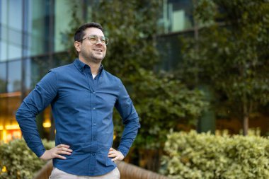 Mavi gömlekli, gözlüklü, gülümseyen, başka tarafa bakan, modern bir binası ve arka planında yeşil ağaçları olan şehir parkında duran kendinden emin bir adam.