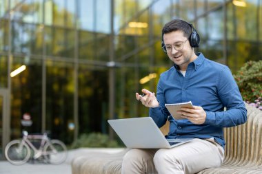 Kulaklık takan ve gözlük takan bir adam dizüstü bilgisayar ve not defteri kullanarak çevrimiçi video görüşmesi yapıyor. Modern bir binanın önündeki bankta oturup esnek uzaktan çalışma ve dijital iletişimi kucaklıyor.