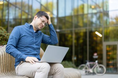 Adam, dışarıda dizüstü bilgisayarla çalışırken, modern bir ofis binasının yanındaki bankta otururken, yorgun ve şiddetli bir baş ağrısı çekiyor.