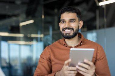 Hintli işadamı elinde dijital tablet tutarken gülümsüyor, çağdaş bir iş yerinde teknoloji, iletişim ve profesyonel başarıyı temsil ediyor.