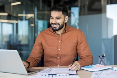 Modern bir ofiste çalışan genç profesyonel bir adam, bilgisayarını kullanırken gülümseyen ve evrak işleriyle uğraşan, üretkenliği ve iş başarısını temsil eden