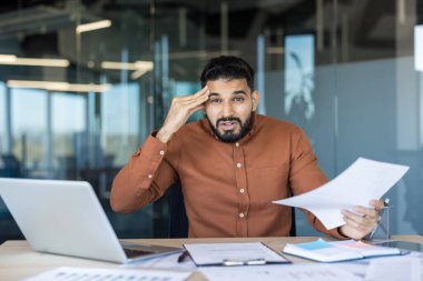 İş adamı stresli ve bunalmış görünüyor, elinde belgelerle ve kafasına dokunarak modern ofis ortamında kağıt ve dizüstü bilgisayarla dolu bir masada oturuyor.