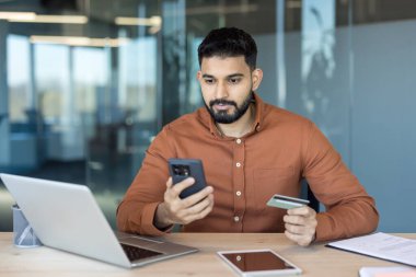 Genç işadamı akıllı telefon ve kredi kartıyla güvenli bir online ödeme yapıyor. Modern bir ofis masasında, mobil bankacılık uygulaması ve kablosuz bağlantı kullanarak ekomünikasyon işlemleri yapıyor.