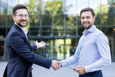 İki gülümseyen iş adamı, bir ortaklık anlaşmasını onaylamak için el sıkışıyor ya da modern bir şirket binasının önünde duran yeni bir meslektaşa hoş geldin diyor.