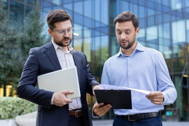 Yeni bir projede işbirliği yapan iki profesyonel işadamı, önemli belgeleri gözden geçiriyorlar ve modern bir ofis binasının önünde dizüstü bilgisayar kullanıyorlar.