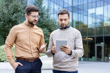 İki işadamı, dijital tablet hakkındaki bilgileri yoğun bir şekilde inceliyor, fikirleri paylaşıyor ve modern bir ofis binası dışında bir soruna çözüm buluyor.