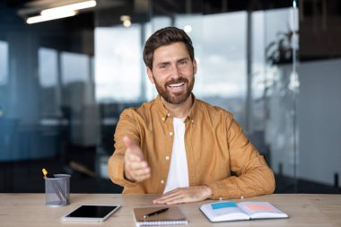 Genç işadamı kameraya bakıyor ve dostça el sıkışmak için elini uzatıyor, modern ofiste masa başında oturuyor, iş görüşmesi kavramı, selamlama, hoş geldin veya ortaklık.
