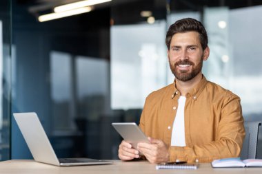 İş adamı kameraya bakıyor, elinde bir dizüstü bilgisayarla masada oturuyor, dijital tablet tutuyor, modern teknolojiyi ve profesyonel çalışma ortamını temsil ediyor.