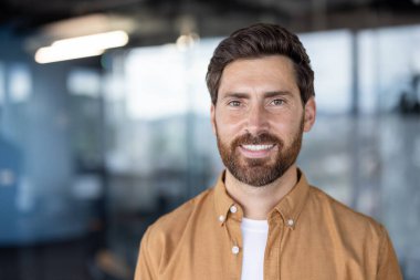 Kameraya gülümseyen, çağdaş ofis ortamında sıradan bir gömlek giyen, profesyonelliği, başarıyı ve pozitif kurumsal kültürü temsil eden kendinden emin sakallı bir adam.