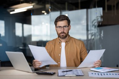 Modern bir ofiste masa başında çalışan kendine güvenen bir işadamı, elinde önemli belgeler, bir dizüstü bilgisayar ve hesap makinesiyle veri analizi, odak ve uzmanlık göstergesi