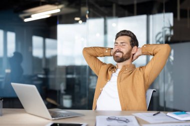 Gülümseyen adam işe ara veriyor, elleri başının arkasında sandalyesine yaslanıyor, modern bir ofiste başarının ve başarının tadını çıkarıyor.