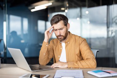 Stresli erkek çalışanın ofis masasında dizüstü bilgisayarla çalışırken yorgunluk ve baş ağrısı çekmesi, mesleki sorunlar ve teslim tarihleriyle uğraşması