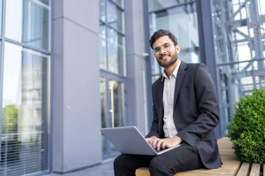 Takım elbiseli genç bir işadamı açık hava bankında oturuyor, dizüstü bilgisayarda gülümsüyor ve daktilo yazıyor, modern ofis binasının dışındaki iş teknolojisine bağlanıyor.