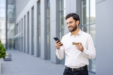 Genç adam dışarıda gülümserken akıllı telefon kullanıyor ve kredi kartı tutuyor, online alışveriş için güvenli cep telefonu ödemesi yapıyor modern, kullanışlı e-ticaret ve dijital bankacılık anı