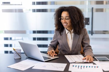 Ofiste oturan Afro-Amerikalı genç iş kadını bir dizüstü bilgisayar ve belgelerle çalışıyor..