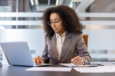 Ciddi ve odaklanmış, takım elbiseli, dizüstü bilgisayar ve belgelerle çalışan Afro-Amerikan kadın..