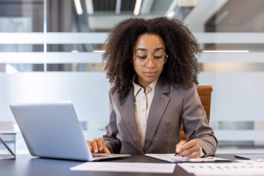 Ciddi ve konsantre olmuş genç bir Afro-Amerikan kadın elinde dizüstü bilgisayarla ofiste oturuyor, belgelerle çalışıyor, veri ve tabloları dolduruyor..