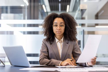 Ciddi ve başarılı bir Afro-Amerikan kadın portresi. Takım elbiseli ve gözlüklü. Ofiste bir masada oturuyor..