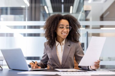 Gülümseyen genç bir Afro-Amerikalı iş kadını ofiste çalışıyor, elinde bir dizüstü bilgisayarla bir masada oturuyor ve elinde belgeler ve faturalar var..