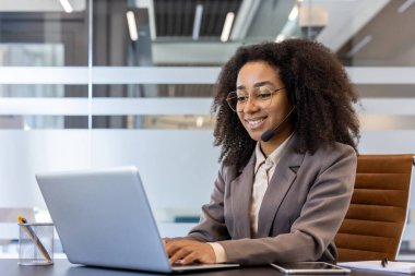 Takım elbiseli, gülümseyen Afro-Amerikan iş kadını ofiste dizüstü bilgisayar üzerinde çalışıyor..