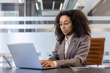 Takım elbiseli ciddi bir Afro-Amerikalı iş kadını ofiste dizüstü bilgisayarla çalışmaya odaklanıyor..