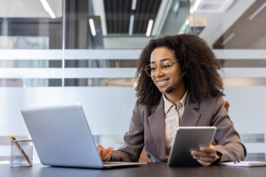 Gülümseyen genç bir Afro-Amerikalı kadın ofisinde oturuyor ve elinde bir dizüstü bilgisayar ve tablet kullanıyor..
