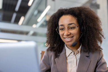 Ofiste dizüstü bilgisayarla çalışan, kulaklık takmış gülümseyen Afro-Amerikan bir iş kadınının yakın plan fotoğrafı..